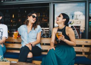 Two women sitting on a bench outside of a brewery, laughing as they sip glasses of beer.